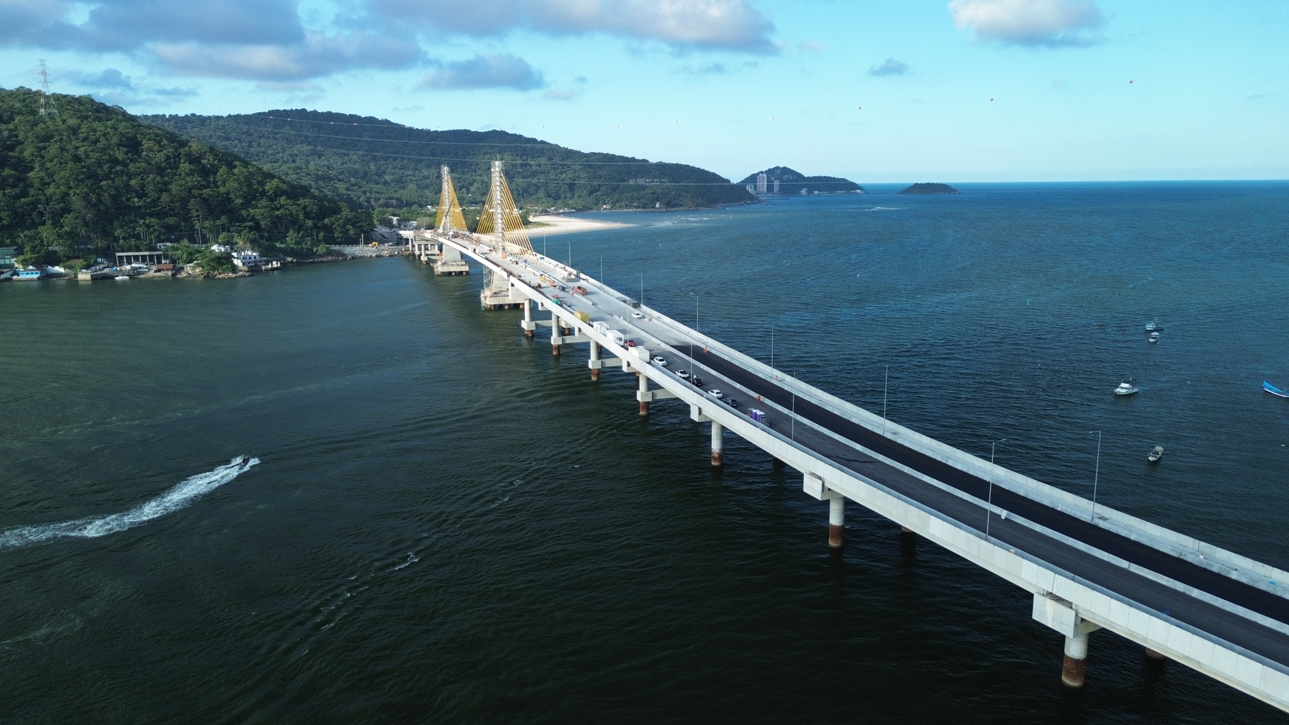 Ponte de Guaratuba vista de cima com pista asfaltada e veículos circulando, sustentada por pilares no mar e cabos estaiados ligados a torres centrais, com embarcações na água ao redor, vegetação costeira próxima e céu com nuvens ao fundo.